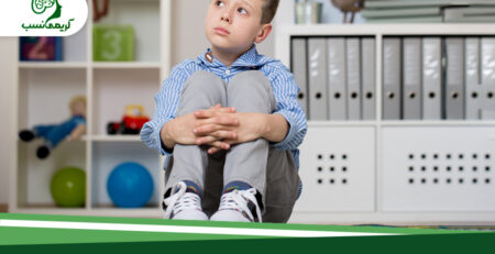 پسربچه اوتیستیک که در اتاقی که پشت سر او یک کتابخانه و یک کمد اسباب بازی قرار دارد نشسته است و زانو های خود را بغل کرده در پایین کادر تیتر علائم اوتیسم در کودکان چیست نوشته شده است